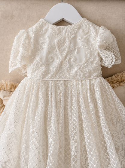 An elegant heirloom gown designed with timeless tradition in mind. Crafted from intricate lace, this gown features pearl and sequence embellishments throughout. It features delicate short sleeves, a softly structured bodice. The back is finished with delicate pearl buttons and a long mikado side belt that can be tied into a bow. The intricate lacework flows throughout the gown, creating beautiful texture and detail from every angle. 