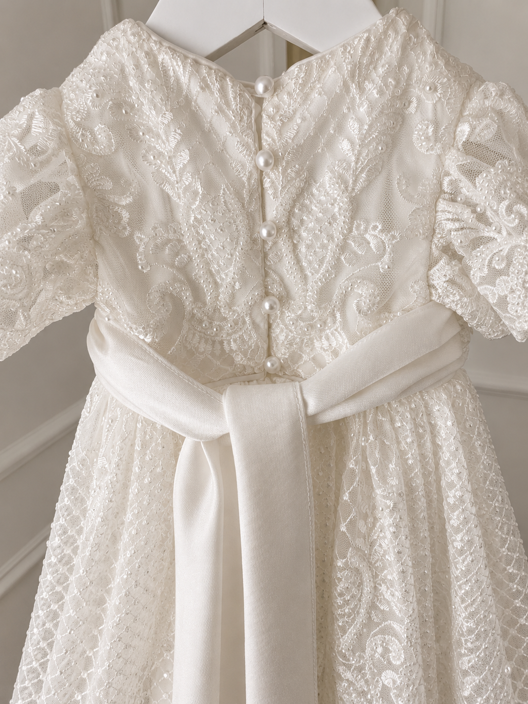 An elegant heirloom gown designed with timeless tradition in mind. Crafted from intricate lace, this gown features pearl and sequence embellishments throughout. It features delicate short sleeves, a softly structured bodice. The back is finished with delicate pearl buttons and a long mikado side belt that can be tied into a bow. The intricate lacework flows throughout the gown, creating beautiful texture and detail from every angle. 