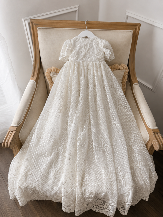 An elegant heirloom gown designed with timeless tradition in mind. Crafted from intricate lace, this gown features pearl and sequence embellishments throughout. It features delicate short sleeves, a softly structured bodice. The back is finished with delicate pearl buttons and a long mikado side belt that can be tied into a bow. The intricate lacework flows throughout the gown, creating beautiful texture and detail from every angle. 
