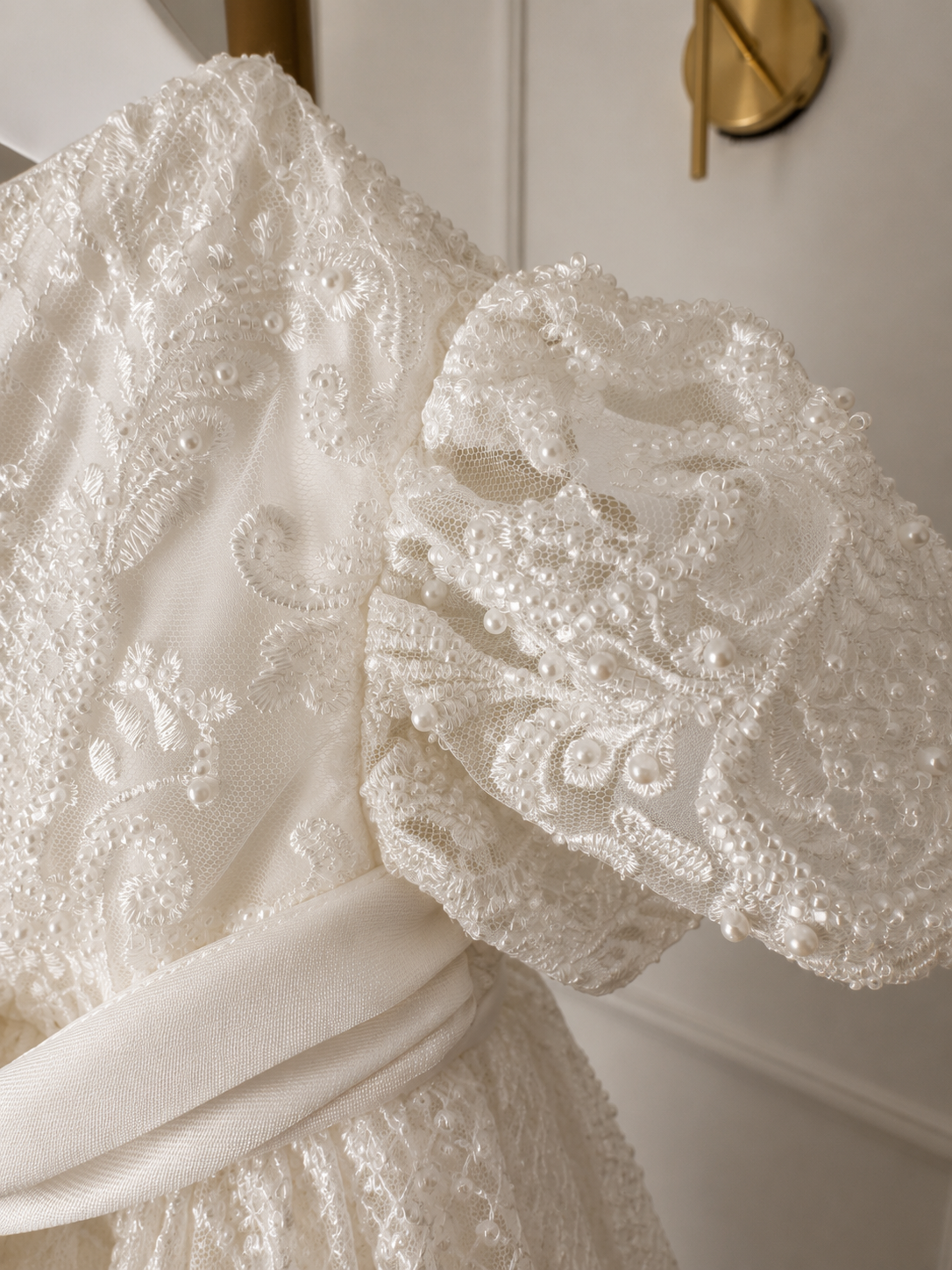 An elegant heirloom gown designed with timeless tradition in mind. Crafted from intricate lace, this gown features pearl and sequence embellishments throughout. It features delicate short sleeves, a softly structured bodice. The back is finished with delicate pearl buttons and a long mikado side belt that can be tied into a bow. The intricate lacework flows throughout the gown, creating beautiful texture and detail from every angle. 