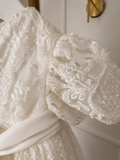 An elegant heirloom gown designed with timeless tradition in mind. Crafted from intricate lace, this gown features pearl and sequence embellishments throughout. It features delicate short sleeves, a softly structured bodice. The back is finished with delicate pearl buttons and a long mikado side belt that can be tied into a bow. The intricate lacework flows throughout the gown, creating beautiful texture and detail from every angle. 