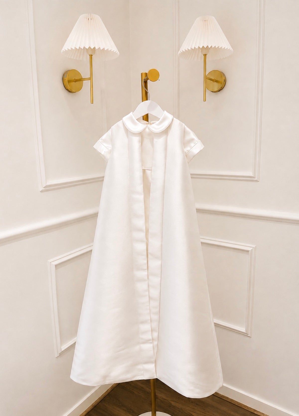 A white short-sleeve dress with a Peter Pan collar hangs on a gold hanger against a white paneled wall with two gold wall sconces.