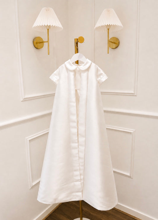 A white short-sleeve dress with a Peter Pan collar hangs on a gold hanger against a white paneled wall with two gold wall sconces.