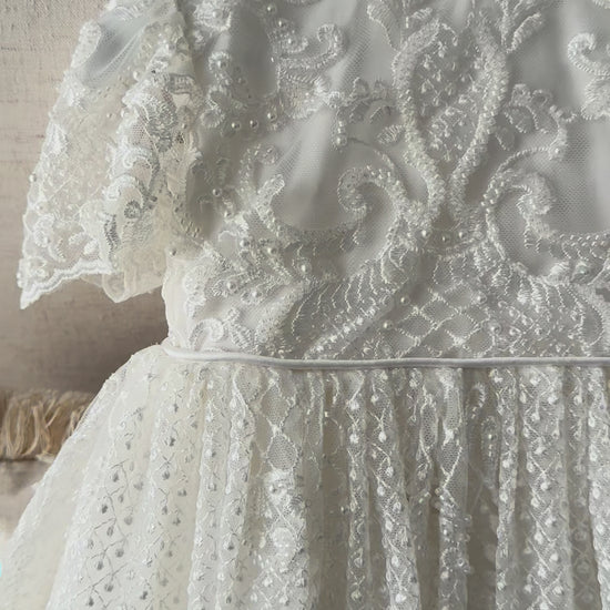 An elegant heirloom gown designed with timeless tradition in mind. Crafted from intricate lace, this gown features pearl and sequence embellishments throughout. It features delicate short sleeves, a softly structured bodice. The back is finished with delicate pearl buttons and a long mikado side belt that can be tied into a bow. The intricate lacework flows throughout the gown, creating beautiful texture and detail from every angle. 