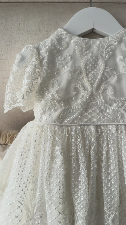 An elegant heirloom gown designed with timeless tradition in mind. Crafted from intricate lace, this gown features pearl and sequence embellishments throughout. It features delicate short sleeves, a softly structured bodice. The back is finished with delicate pearl buttons and a long mikado side belt that can be tied into a bow. The intricate lacework flows throughout the gown, creating beautiful texture and detail from every angle. 