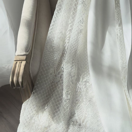 An elegant heirloom gown designed with timeless tradition in mind. Crafted from intricate lace, this gown features pearl and sequence embellishments throughout. It features delicate short sleeves, a softly structured bodice. The back is finished with delicate pearl buttons and a long mikado side belt that can be tied into a bow. The intricate lacework flows throughout the gown, creating beautiful texture and detail from every angle. 
