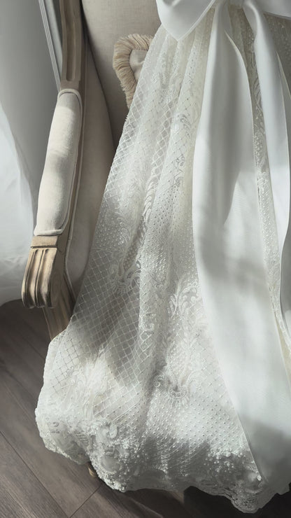 An elegant heirloom gown designed with timeless tradition in mind. Crafted from intricate lace, this gown features pearl and sequence embellishments throughout. It features delicate short sleeves, a softly structured bodice. The back is finished with delicate pearl buttons and a long mikado side belt that can be tied into a bow. The intricate lacework flows throughout the gown, creating beautiful texture and detail from every angle. 