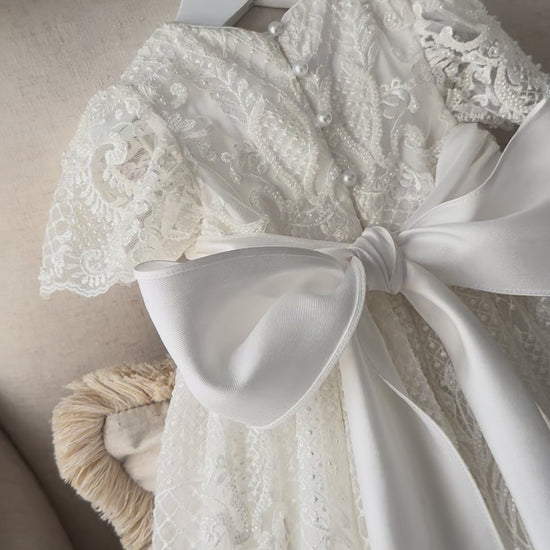 An elegant heirloom gown designed with timeless tradition in mind. Crafted from intricate lace, this gown features pearl and sequence embellishments throughout. It features delicate short sleeves, a softly structured bodice. The back is finished with delicate pearl buttons and a long mikado side belt that can be tied into a bow. The intricate lacework flows throughout the gown, creating beautiful texture and detail from every angle. 