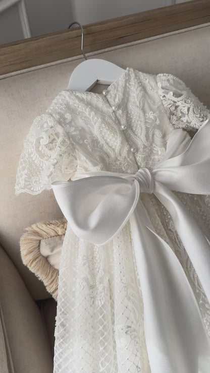 An elegant heirloom gown designed with timeless tradition in mind. Crafted from intricate lace, this gown features pearl and sequence embellishments throughout. It features delicate short sleeves, a softly structured bodice. The back is finished with delicate pearl buttons and a long mikado side belt that can be tied into a bow. The intricate lacework flows throughout the gown, creating beautiful texture and detail from every angle. 