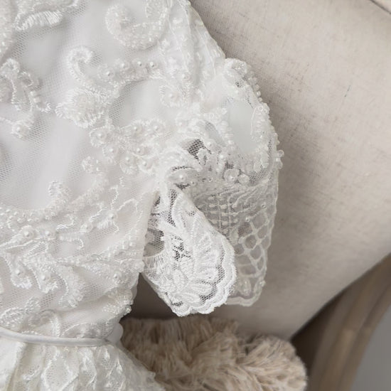 An elegant heirloom gown designed with timeless tradition in mind. Crafted from intricate lace, this gown features pearl and sequence embellishments throughout. It features delicate short sleeves, a softly structured bodice. The back is finished with delicate pearl buttons and a long mikado side belt that can be tied into a bow. The intricate lacework flows throughout the gown, creating beautiful texture and detail from every angle. 