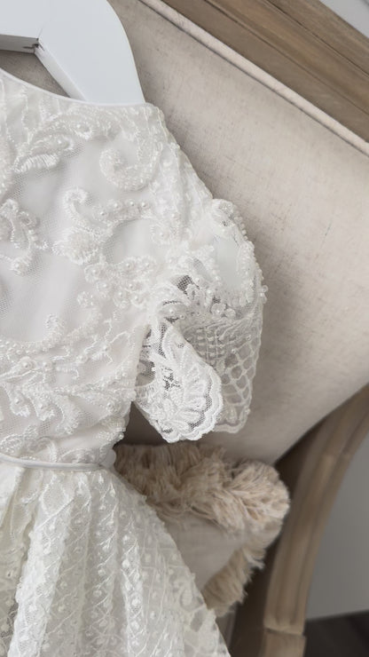 An elegant heirloom gown designed with timeless tradition in mind. Crafted from intricate lace, this gown features pearl and sequence embellishments throughout. It features delicate short sleeves, a softly structured bodice. The back is finished with delicate pearl buttons and a long mikado side belt that can be tied into a bow. The intricate lacework flows throughout the gown, creating beautiful texture and detail from every angle. 