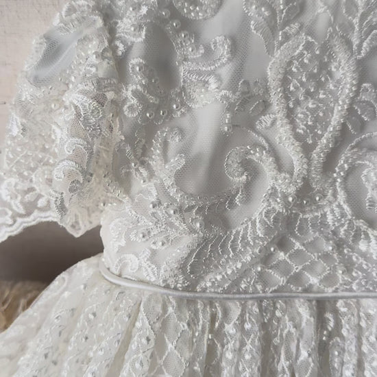 An elegant heirloom gown designed with timeless tradition in mind. Crafted from intricate lace, this gown features pearl and sequence embellishments throughout. It features delicate short sleeves, a softly structured bodice. The back is finished with delicate pearl buttons and a long mikado side belt that can be tied into a bow. The intricate lacework flows throughout the gown, creating beautiful texture and detail from every angle. 