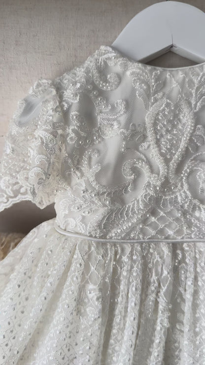 An elegant heirloom gown designed with timeless tradition in mind. Crafted from intricate lace, this gown features pearl and sequence embellishments throughout. It features delicate short sleeves, a softly structured bodice. The back is finished with delicate pearl buttons and a long mikado side belt that can be tied into a bow. The intricate lacework flows throughout the gown, creating beautiful texture and detail from every angle. 